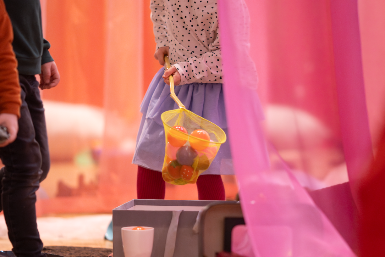 Child in blue skirt holding a yellow fishing net containing coloured balls