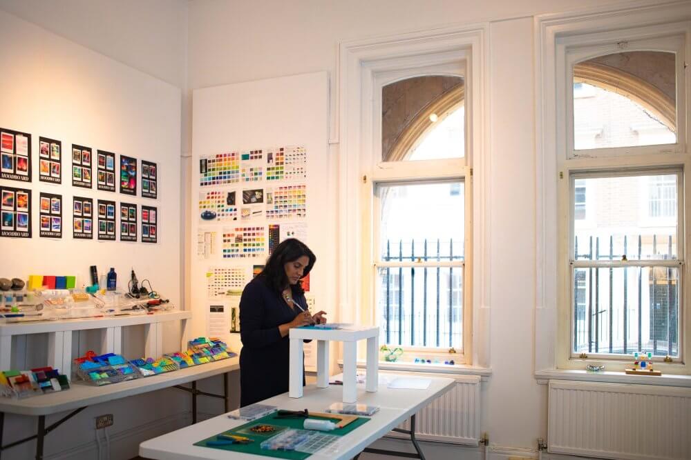 Madiha Zaidi working in her bright glass art studio, surrounded by colour charts, mood boards, and vibrant fused glass, as she prepares a new jewellery piece on a raised work surface.
