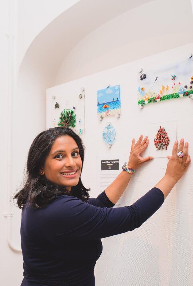 Madiha Zaidi smiling while holding her latest fused glass artwork featuring a log fire made from red crinkled glass, displayed on a white gallery wall. Surrounding pieces include a beach scene, a tree, and a meadow - each part of her nature’s elements theme.