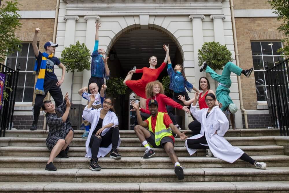 11 dancers on the front steps of a public building. Some sit, some pose with dance moves. They wear costumes such as a high-vis vest, a football scarf and a doctor’s white coat.