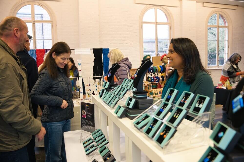 Madiha at the Christmas Shopping Weekend at Orleans House Gallery. Madiha stands to the right behind her stall, which is made up of jewellery in blue boxes. Two customers look at her stall. In the background, people visit other stalls.