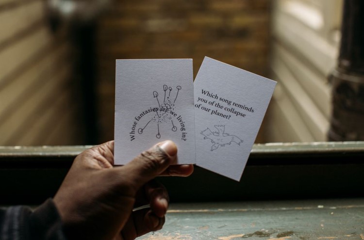 A hand holds two cards the size of playing cards. One reads: Whose fantasies are we living in? The other reads: Which song reminds you of the collapse of our planet?
