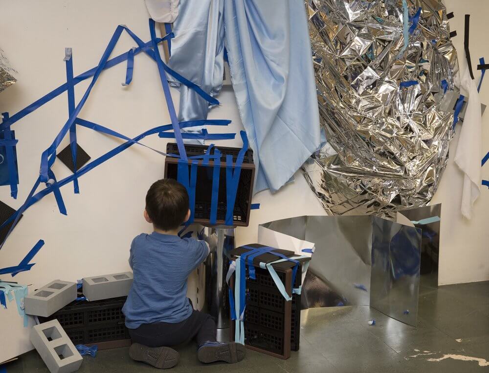 a child explores materials, including fabric, blue tape and silver foil.