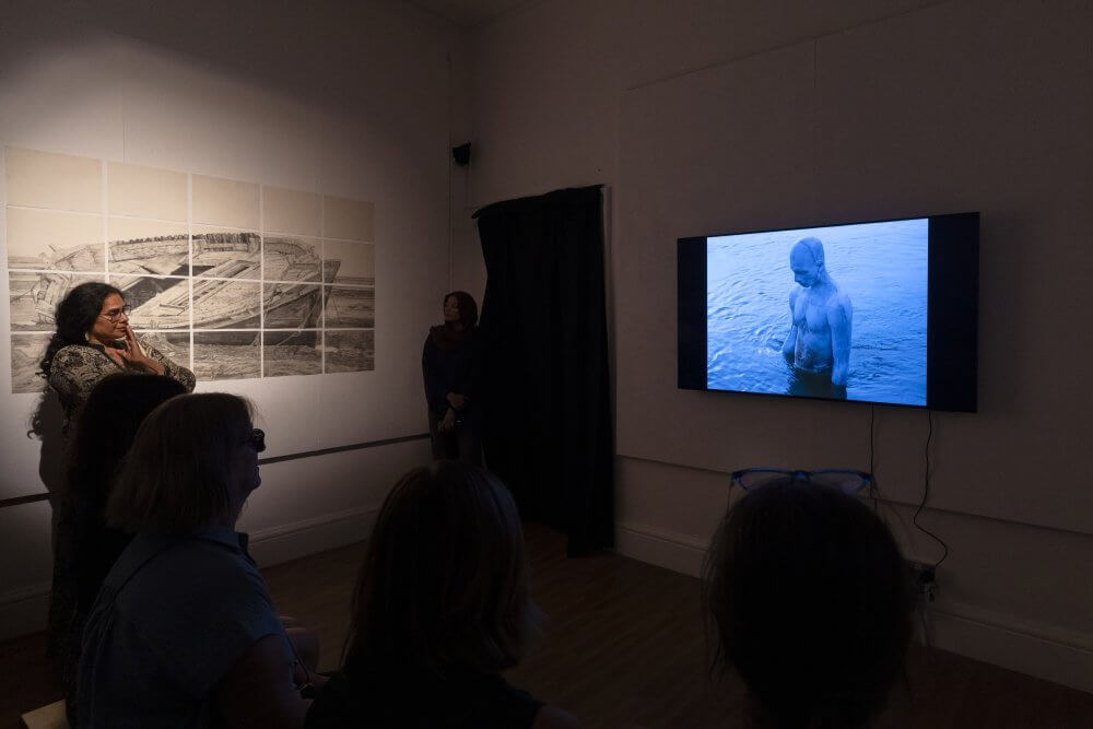 People gather in an exhibition space to watch the River Sublime film. The Broken Boat drawing is on the wall near the film.