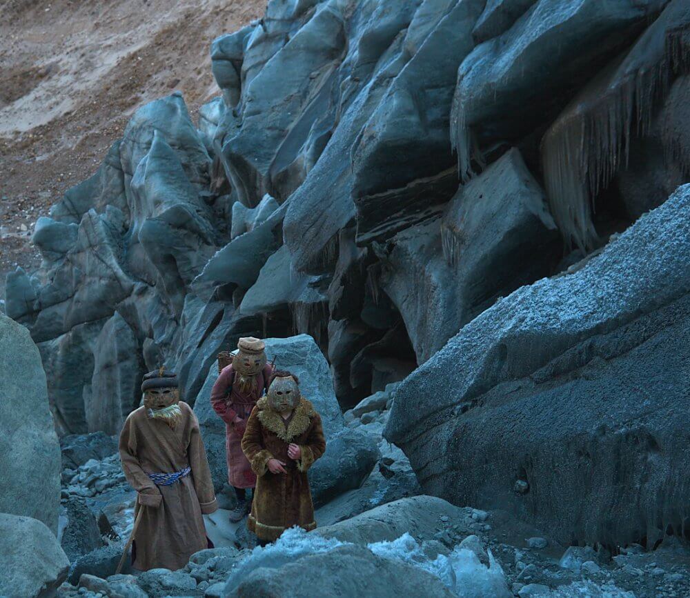 Three people wearing wooden masks walk up a rocky mountainside. At least one carries a wicker basket on their back