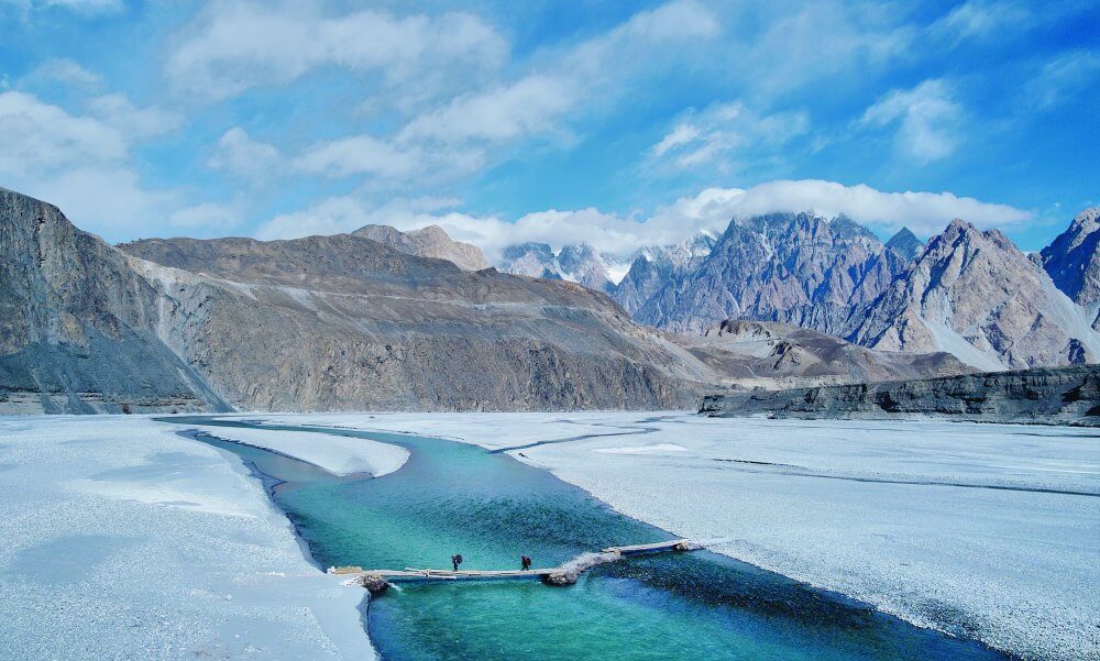 Two people cross a blue river via a footbridge. A rocky mountain landscape is in the background.