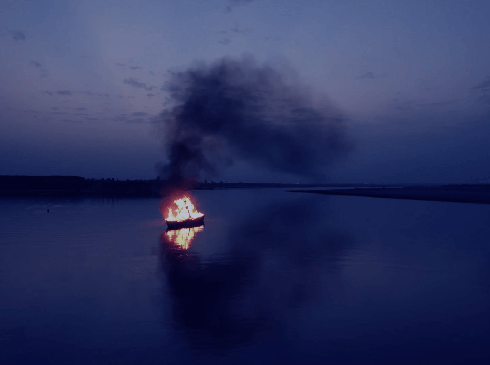 a fishing boat burns in flames on a still, empty river at night