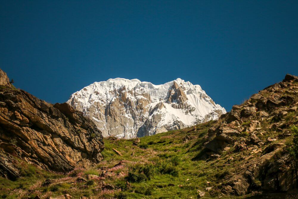 A snow-capped mountain peak in the Hunza Valley with a rocky landscape in the foreground. Grass grows on the rocky foreground.