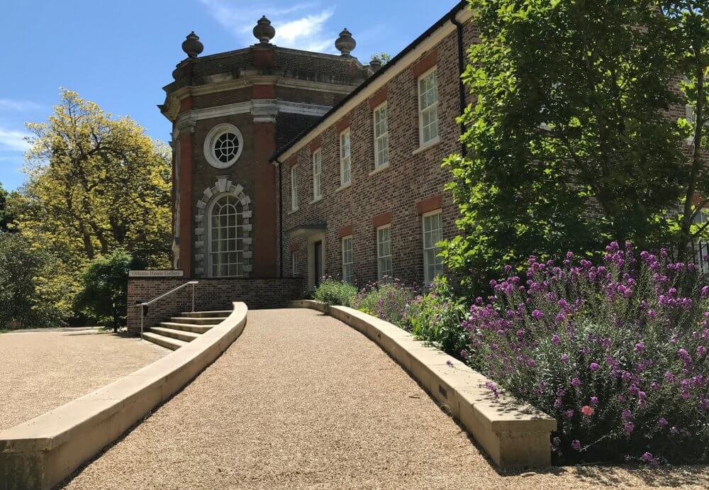 Gravel pathway lined with trees and flowers. Leading to a historic red brick building.